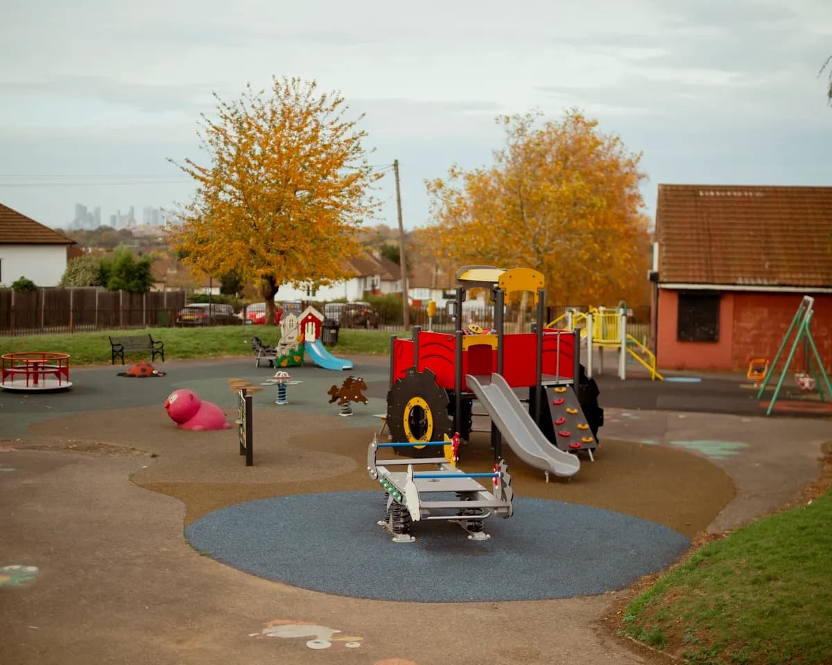 A colorful playground with swings, slides, and other play structures, surrounded by autumn trees and houses, with a city skyline in the distance.