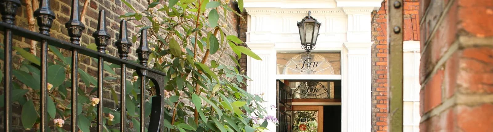 A black iron fence and green foliage in the foreground obscure part of a white ornate doorway with a glass lantern and the word "San" visible through the glass.